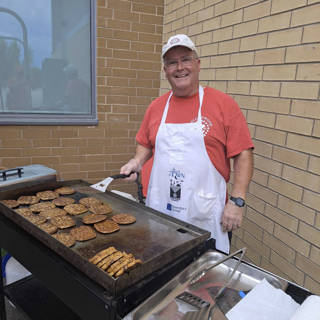 Man in an apron at a barbecue flipping burgers with a Kin Canada tshirt 