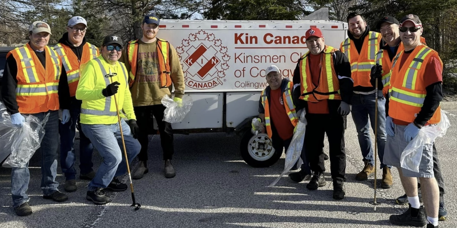 A group of people wearing safety vests looking at the camera smiling with a kin Canada sign on a trailer behind them